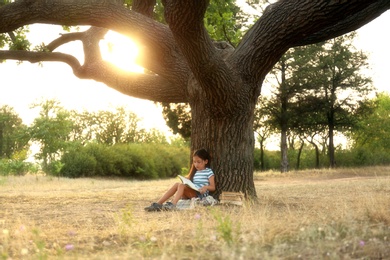 Photo of Cute little girl reading book near tree in park