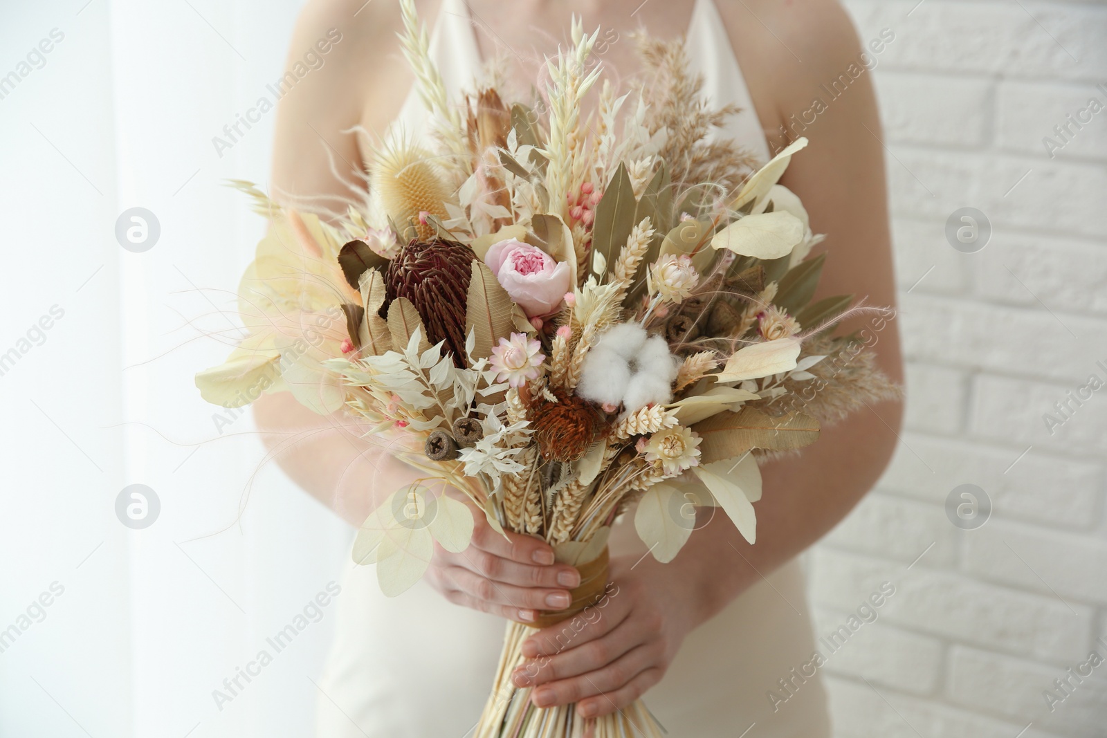 Photo of Bride holding beautiful dried flower bouquet near window at home, closeup