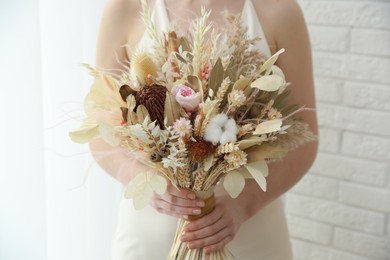 Bride holding beautiful dried flower bouquet near window at home, closeup