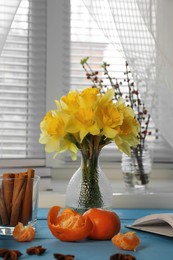 Photo of Beautiful yellow daffodils in vase, tangerines and cinnamon on light blue table