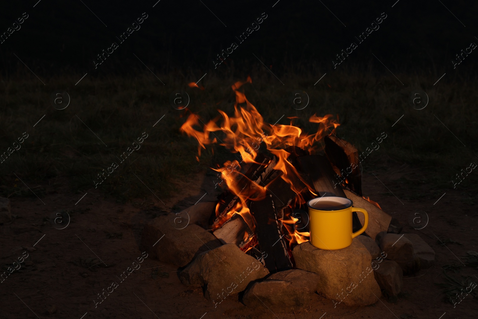 Photo of Metal mug with drink near beautiful bonfire outdoors at night. Camping season