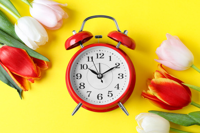 Red alarm clock and spring flowers on yellow background, flat lay. Time change