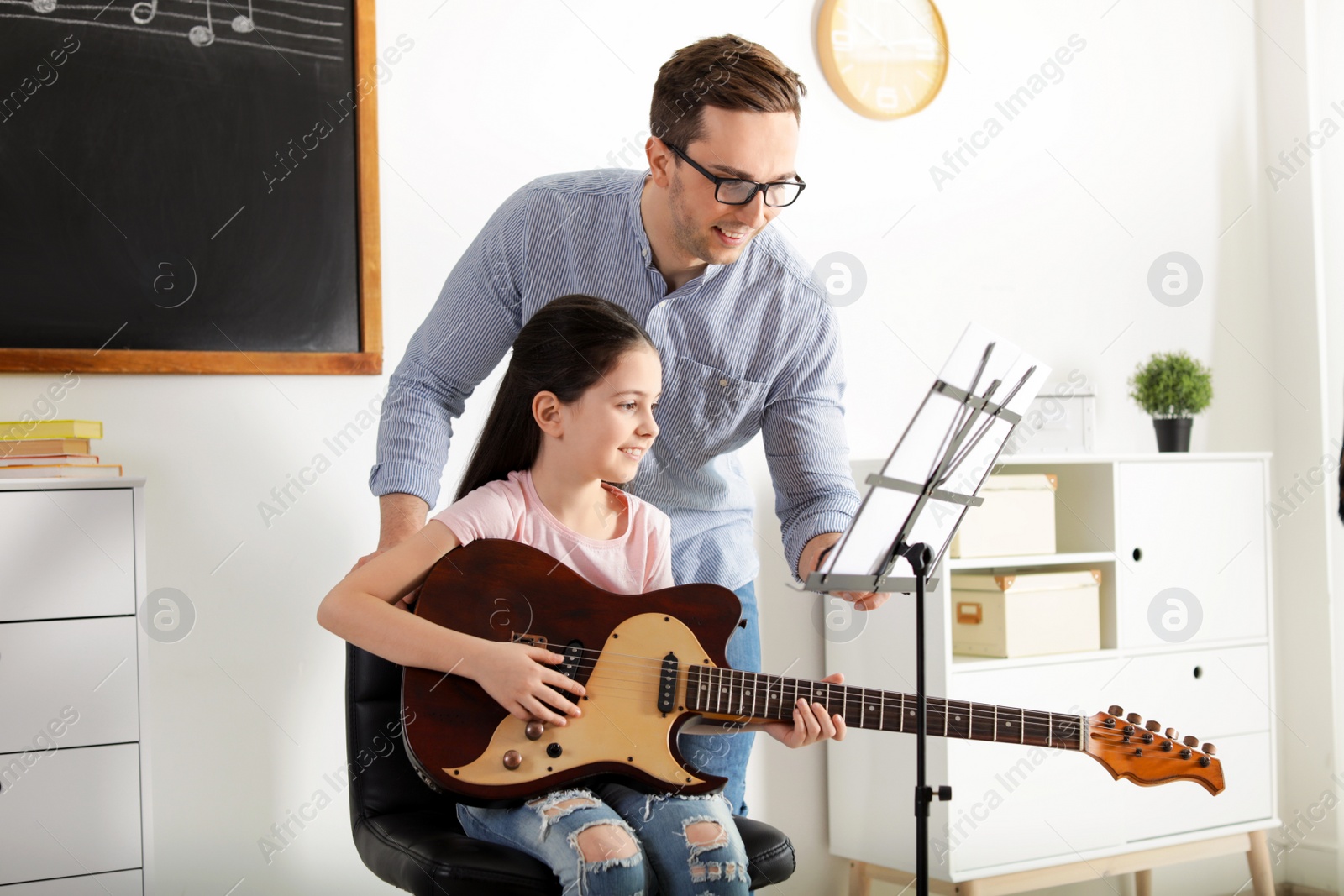 Photo of Little girl playing guitar with her teacher at music lesson. Learning notes