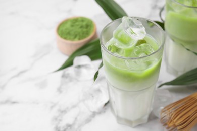 Glasses of tasty iced matcha latte, bamboo whisk and leaves on white marble table, closeup. Space for text