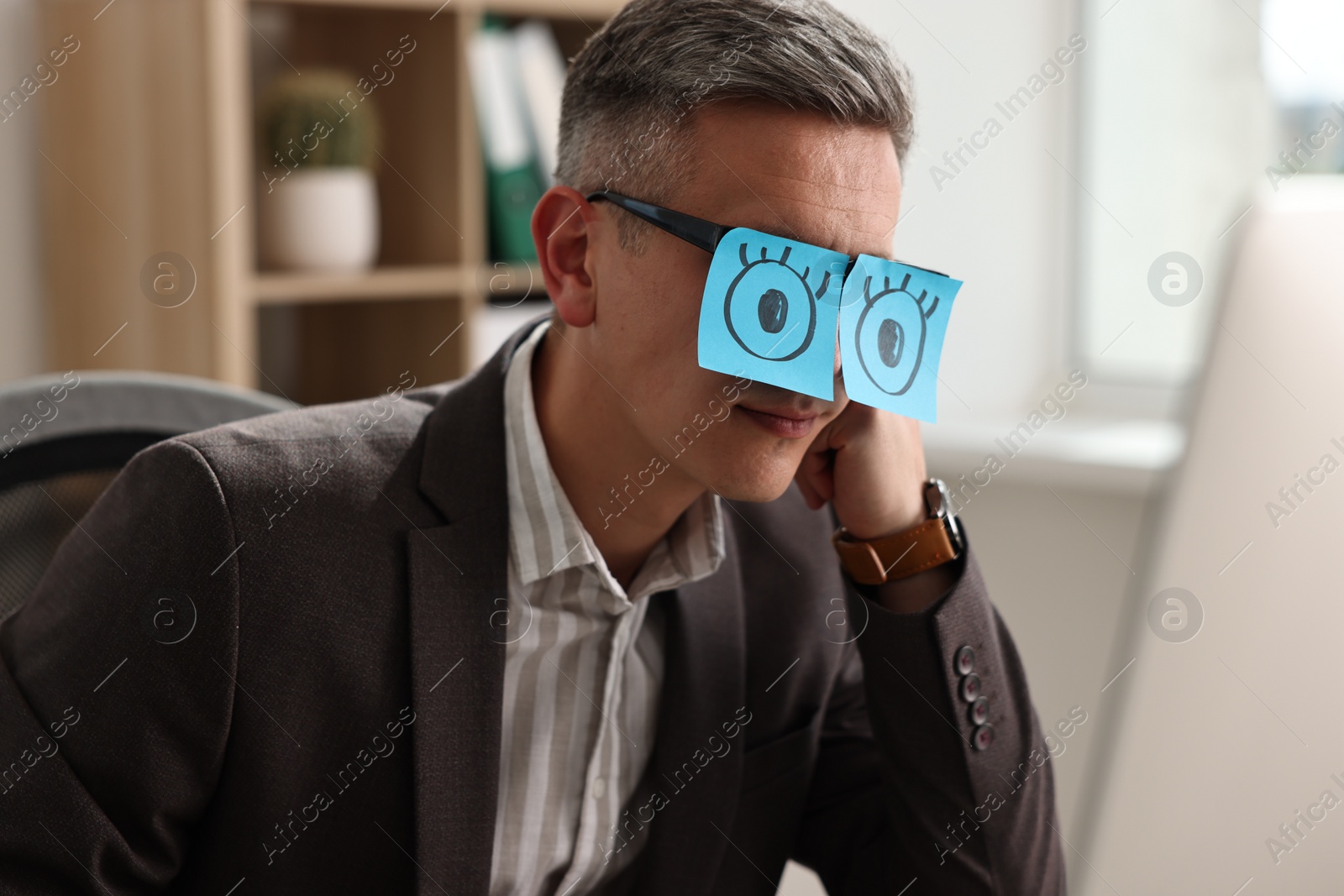 Photo of Man with fake eyes painted on sticky notes snoozing at workplace in office