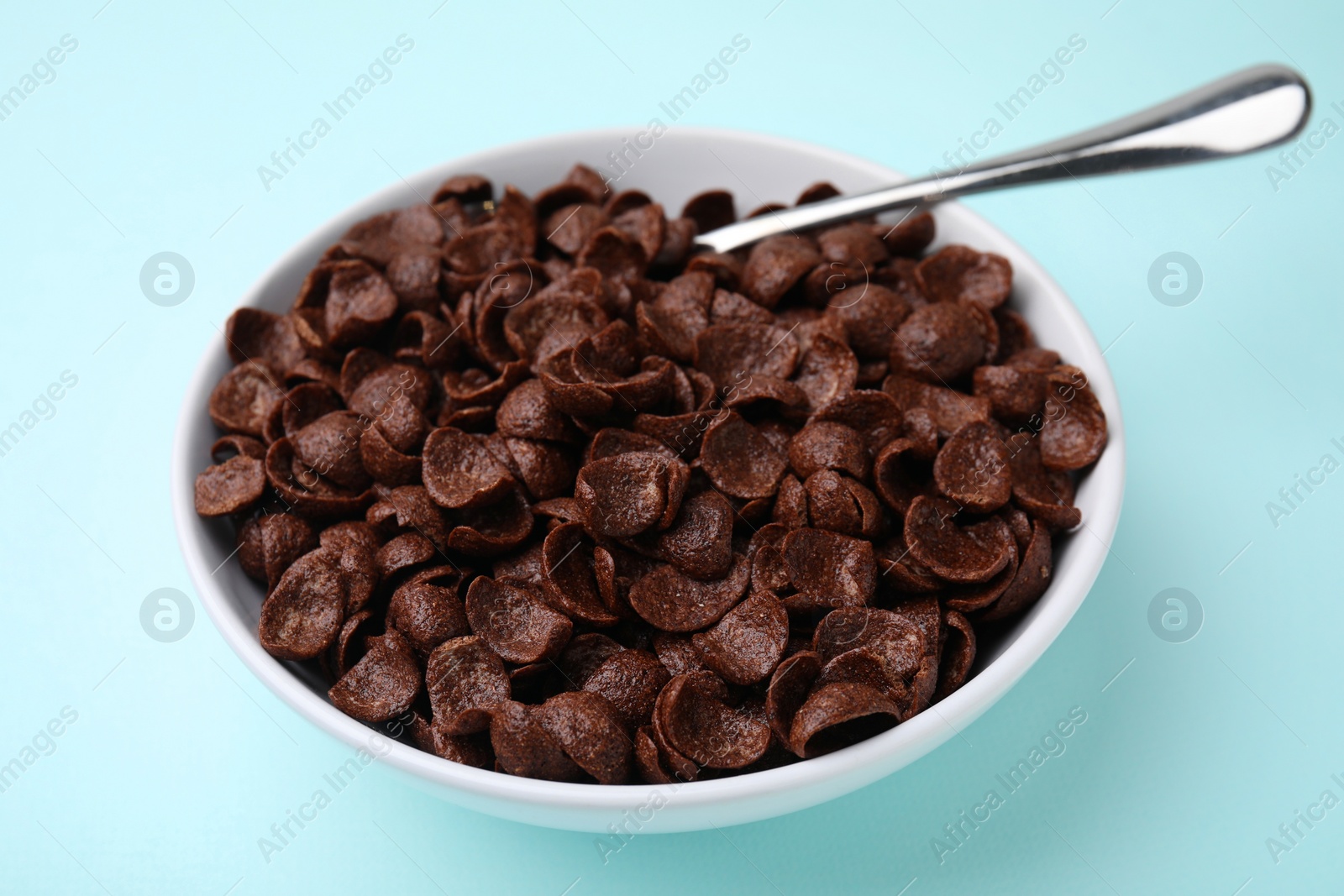 Photo of Breakfast cereal. Chocolate corn flakes in bowl and spoon on light blue table, closeup