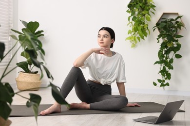 Beautiful girl sitting on yoga mat near laptop in studio