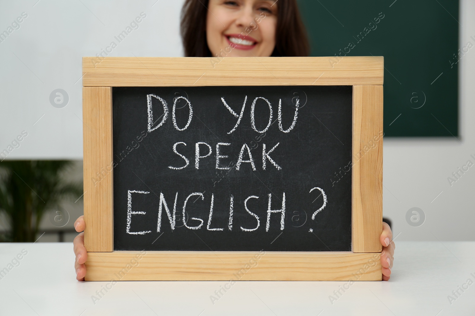 Photo of Teacher holding small chalkboard with inscription Do You Speak English? at table in classroom