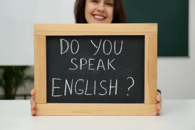 Teacher holding small chalkboard with inscription Do You Speak English? at table in classroom