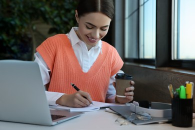 Young female student with laptop drinking coffee while studying at table in cafe