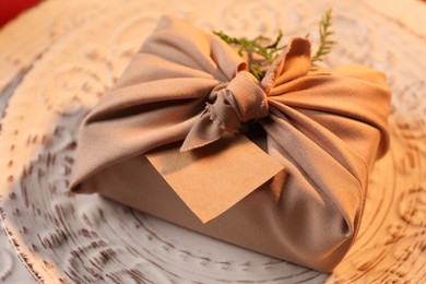 Photo of Furoshiki technique. Gift packed in beige fabric with blank card on plate, closeup