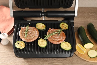 Photo of Electric grill with homemade sausages, zucchini and rosemary on wooden table