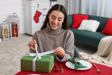 Photo of Happy young woman opening Christmas gift at served table in room