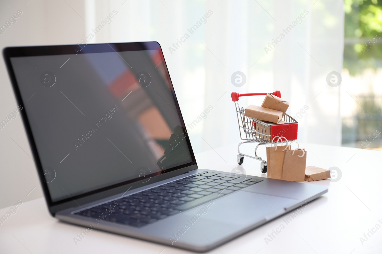 Photo of Internet shopping. Modern laptop, small cart with boxes and bags on table indoors
