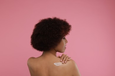 Beautiful young woman applying body cream onto back on pink background