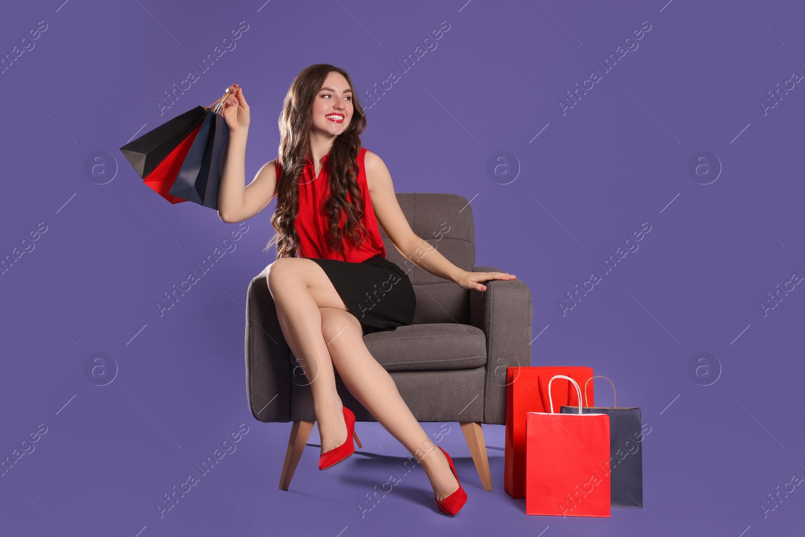 Photo of Happy woman holding paper shopping bags on armchair against purple background