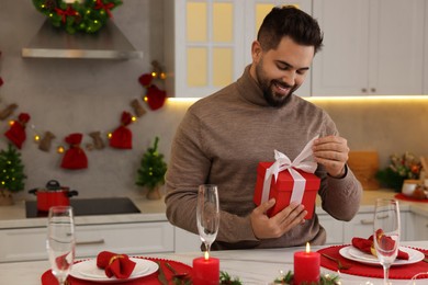 Photo of Happy young man opening Christmas gift at table in kitchen