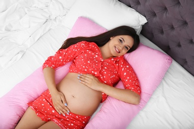 Beautiful pregnant woman lying with maternity pillow on bed at home, top view