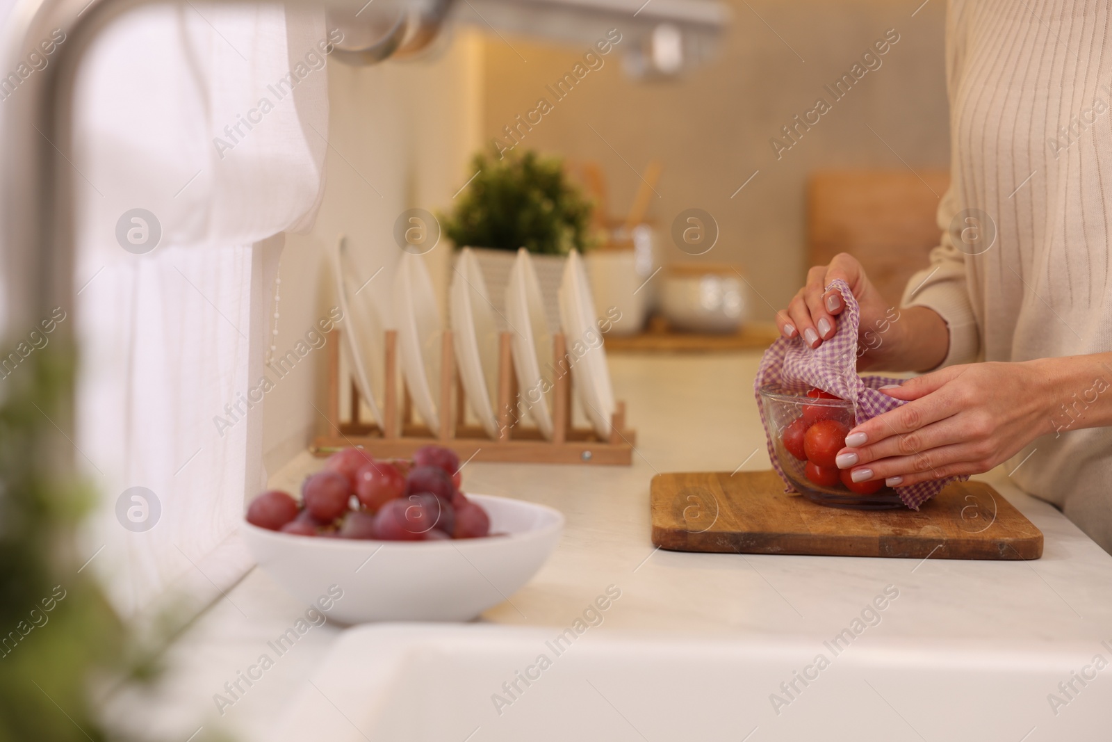 Photo of Woman packing bowl of fresh tomatoes into beeswax food wrap at white countertop in kitchen, closeup