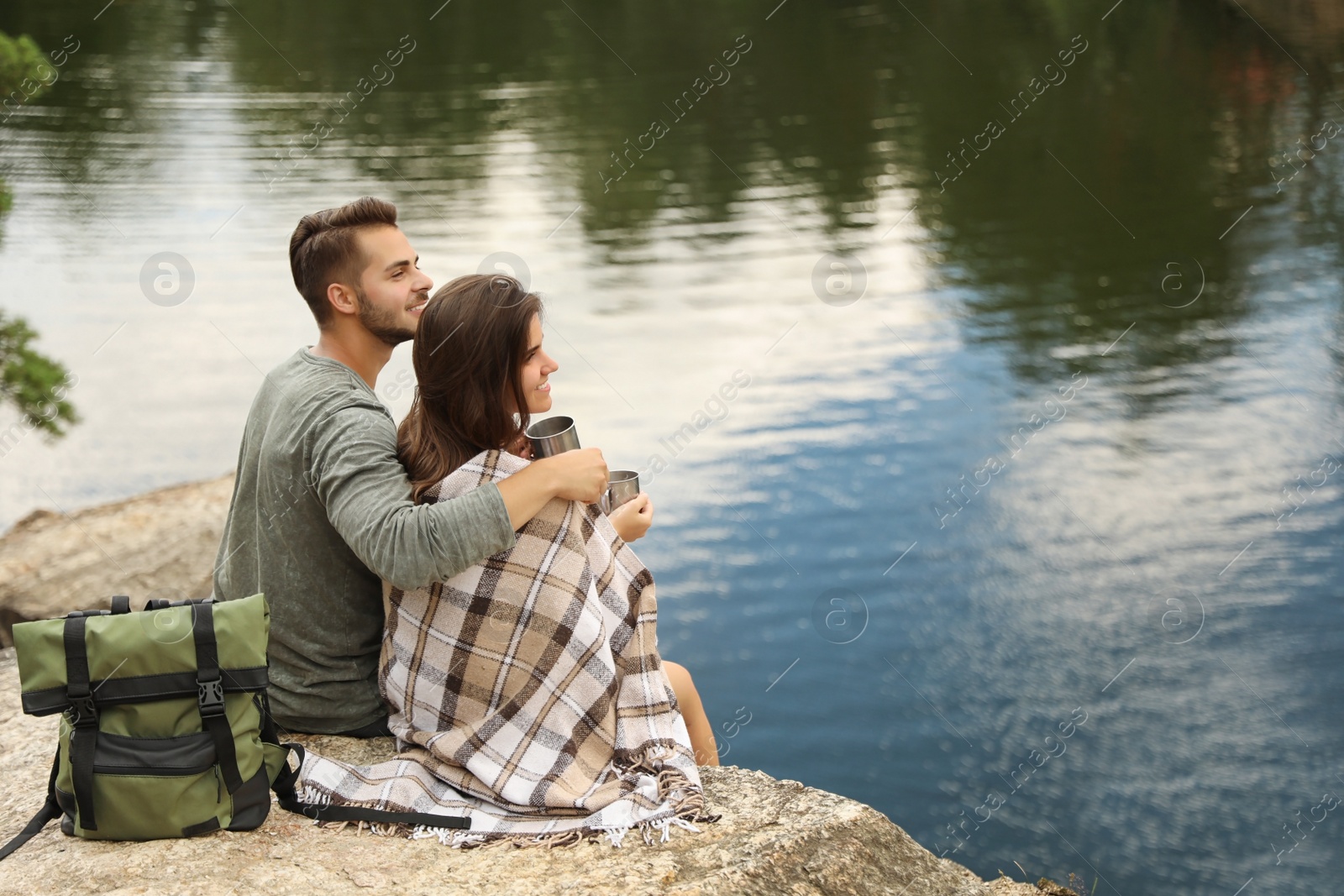 Photo of Cute couple with mugs and plaid near lake. Camping season