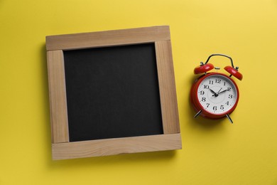 Photo of Blank chalkboard with alarm clock on yellow background, flat lay. Space for text
