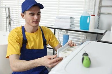 Photo of Young plumber pulling detergent drawer out of washing machine in bathroom