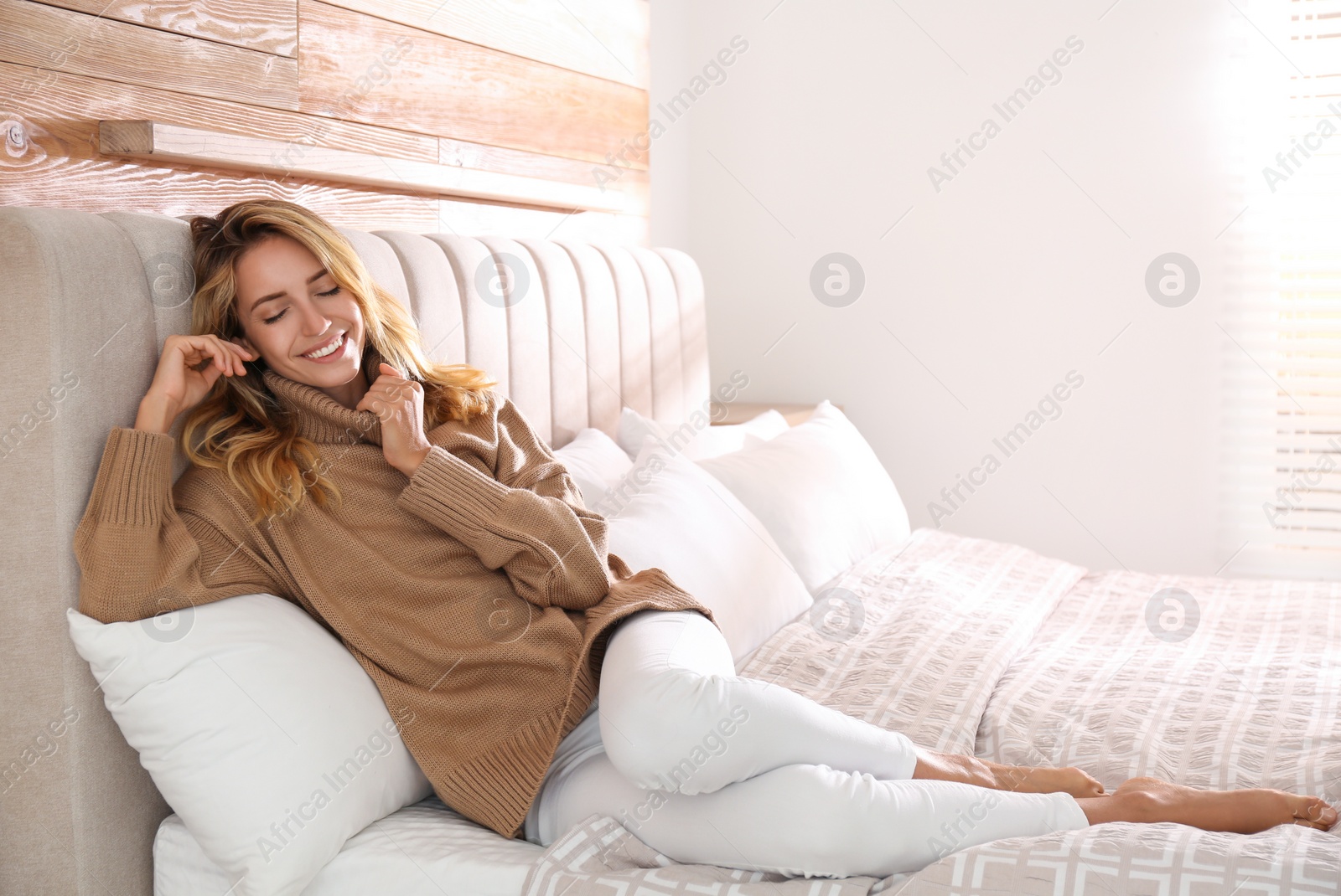 Photo of Beautiful young woman wearing warm brown sweater on bed at home. Space for text