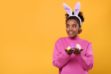 Photo of Happy African American woman in bunny ears headband holding Easter eggs on orange background, space for text