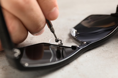 Photo of Handyman repairing sunglasses with screwdriver at grey table, closeup