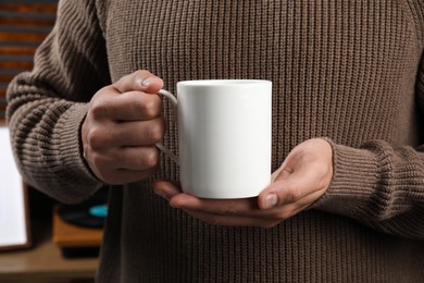 Photo of Man holding white mug indoors, closeup. Mockup for design