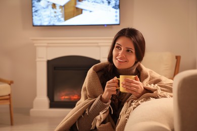 Photo of Young woman with cup of tea relaxing near fireplace at home