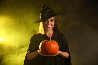 Young woman wearing witch costume with pumpkin in smoke cloud on dark background. Halloween party