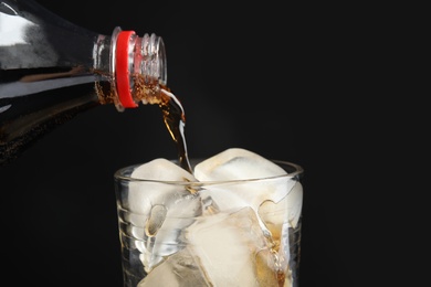 Photo of Pouring refreshing cola from bottle into glass with ice cubes on black background, closeup