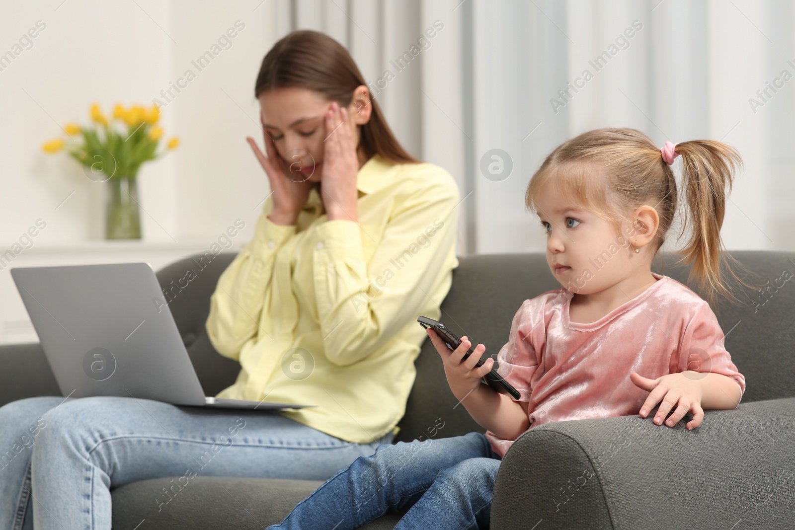 Photo of Tired mother working remotely on laptop while her daughter playing with smartphone at home, selective focus