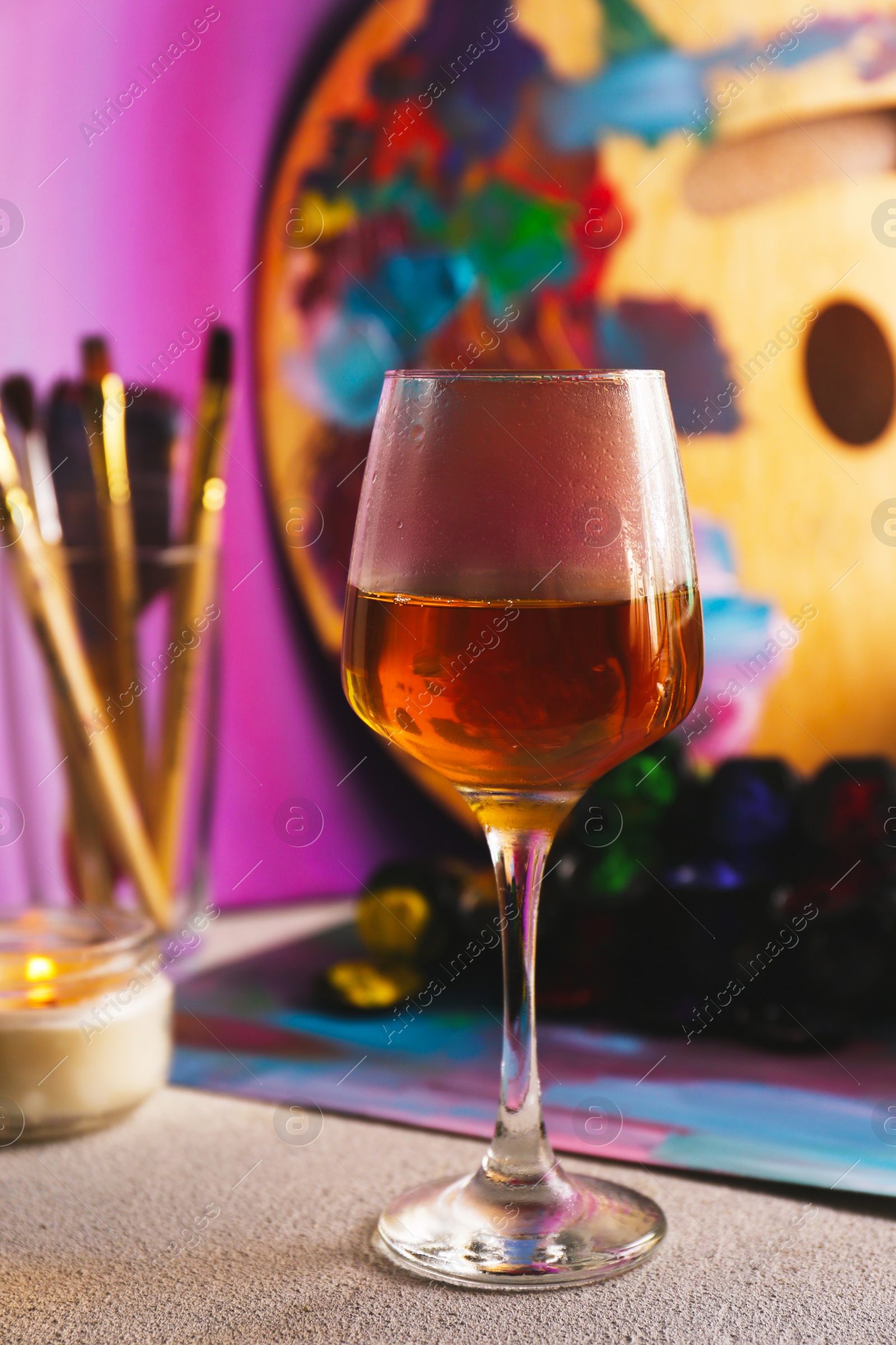 Photo of Glass of tasty wine, brushes with colorful paints and burning candle on light gray table