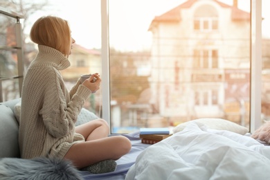 Photo of Beautiful young woman in knitted sweater sitting with cup and looking at window in room. Winter atmosphere