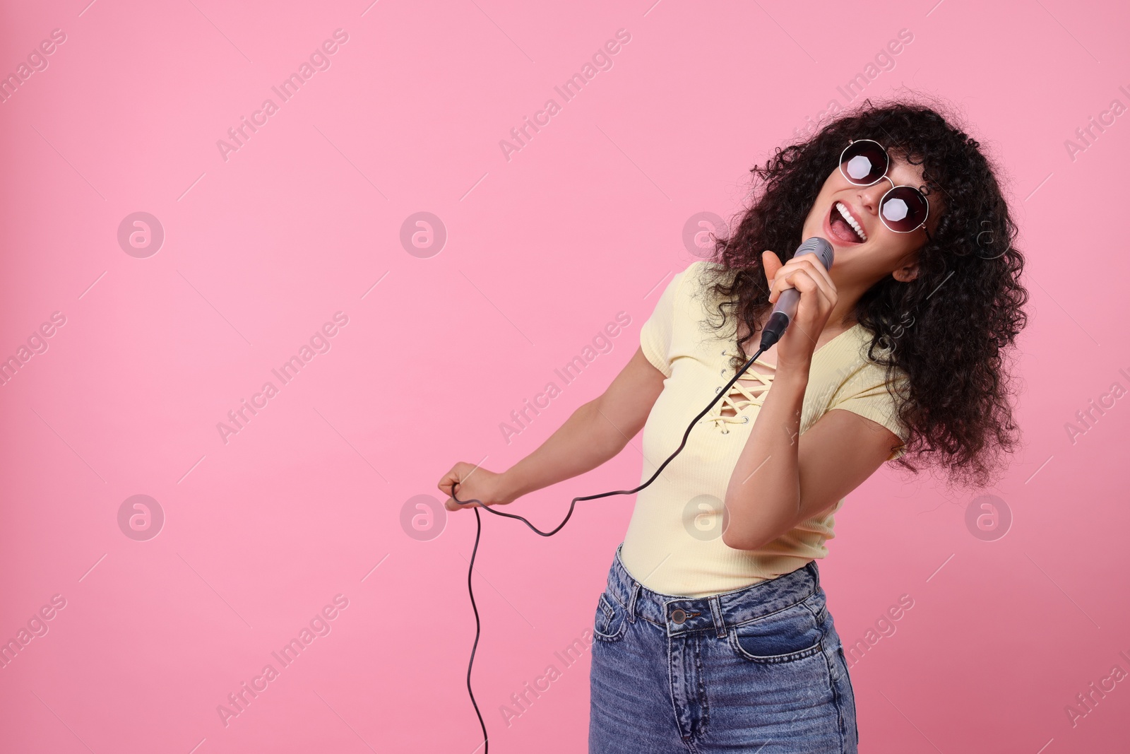 Photo of Beautiful young woman with microphone and sunglasses singing on pink background. Space for text