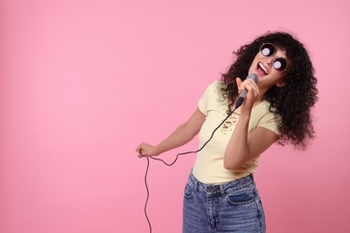 Beautiful young woman with microphone and sunglasses singing on pink background. Space for text