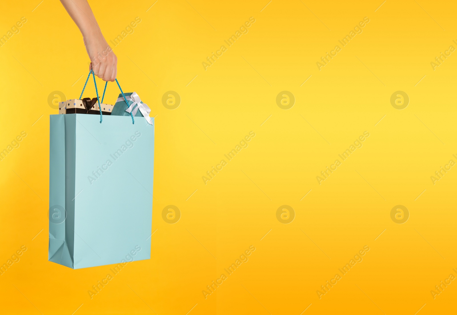 Photo of Woman holding paper shopping bag full of gift boxes on yellow background, closeup