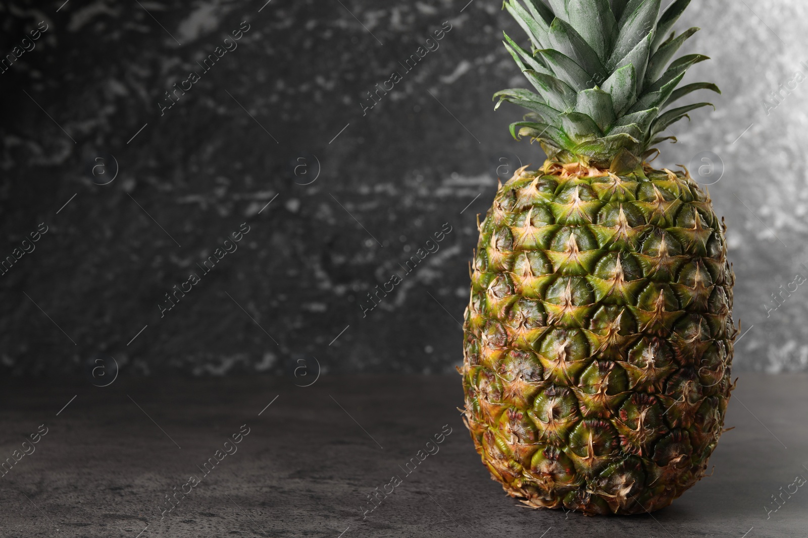 Photo of Whole ripe pineapple on grey table near black wall. Space for text