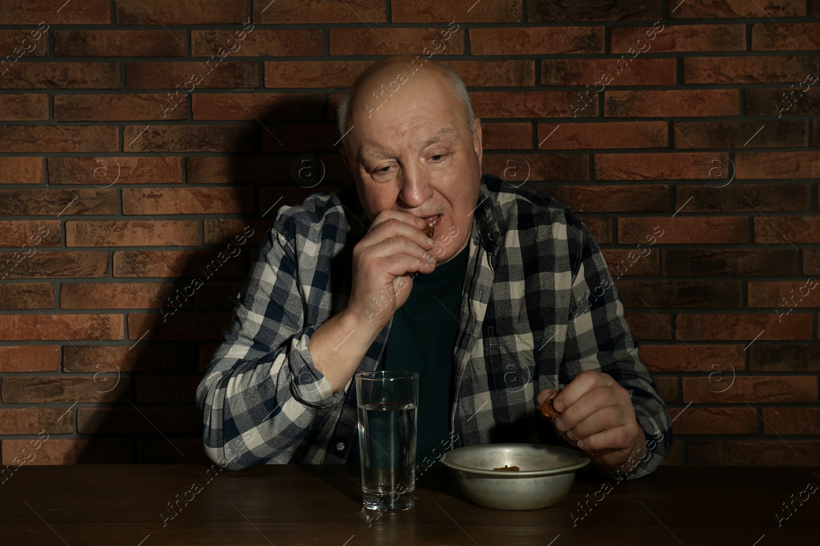 Photo of Poor senior man eating bread at table near brick wall