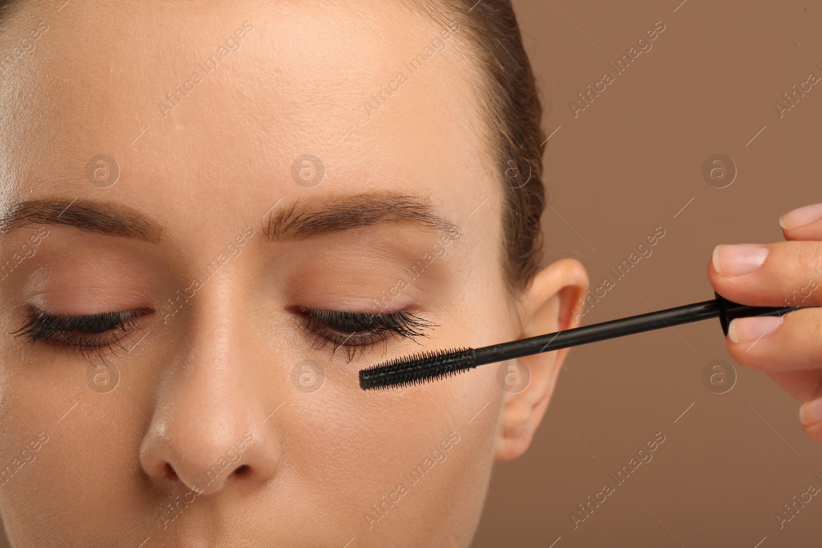 Photo of Woman applying mascara onto eyelashes against light brown background, closeup