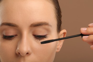 Woman applying mascara onto eyelashes against light brown background, closeup