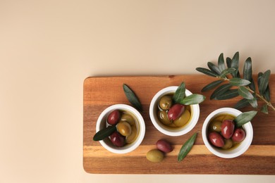 Bowls with different ripe olives and leaves on beige background, flat lay. Space for text