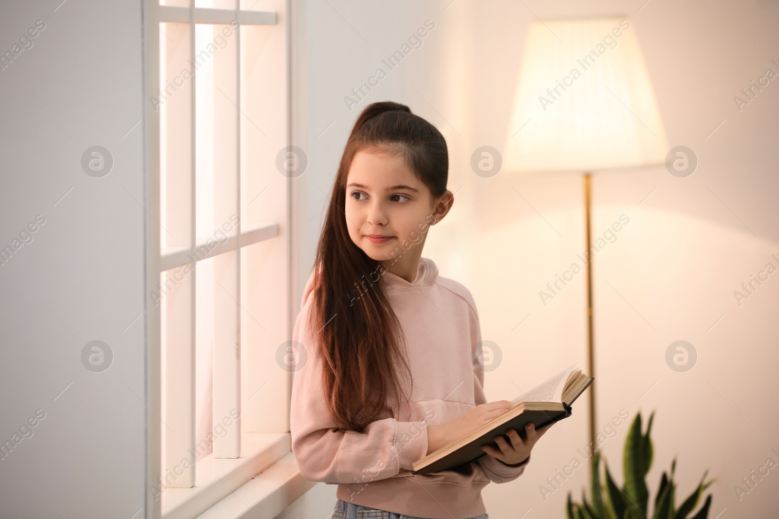 Photo of Cute little girl reading book near window at home