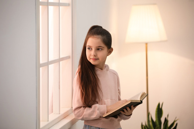 Cute little girl reading book near window at home