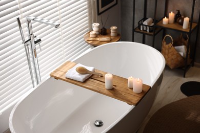 Photo of Wooden tray with burning candles, towel and brush on bathtub in bathroom