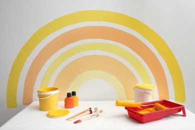 Photo of Different decorator's tools on white table near wall with painted rainbow indoors