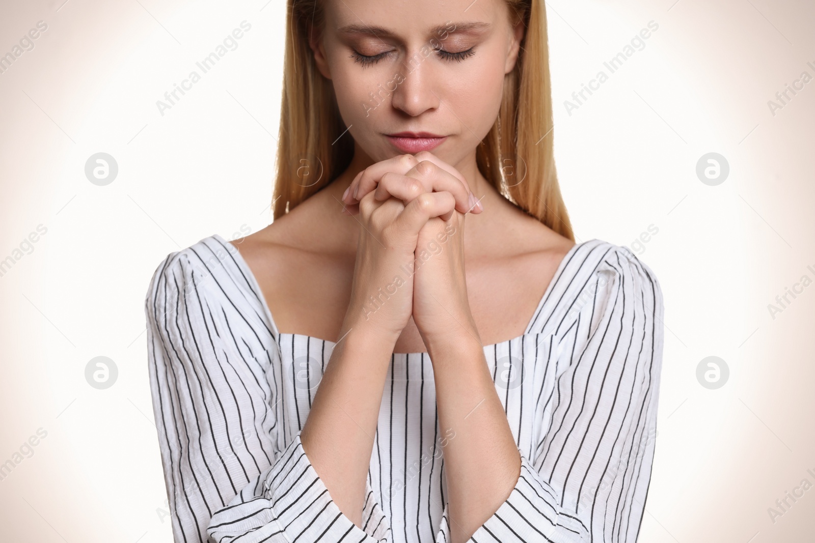 Photo of Religious young woman with clasped hands praying against light background, closeup