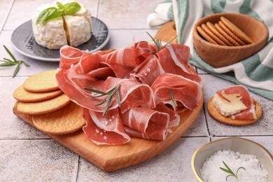 Slices of tasty cured ham, crackers and rosemary on tiled table, closeup